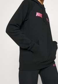 Svart hoodie med rosa Nike-logotyp på bröstet och huvan. Har en framficka, ribbade ärmslut och nederkant för en snug passform. Mjuk tygtextur.