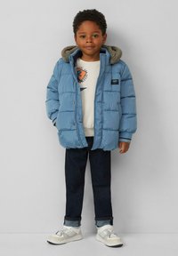 Blauwe pufferjack met een capuchon, geribde manchetten en een klein logo. Gedragen over een licht trui en donkere spijkerbroek met opgerolde manchetten en witte sneakers.