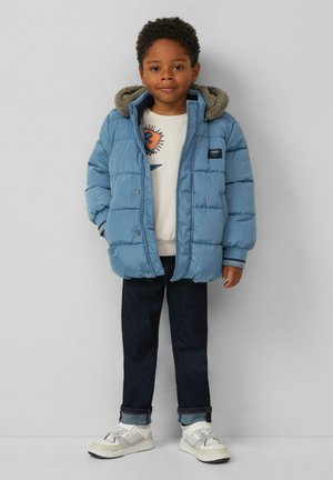 Blaues Daunenjacke mit Kapuze, gerippten Bündchen und kleinem Logo. Getragen über einem hellen Pullover und dunklen Jeans mit aufgeschlagenen Säumen sowie weißen Sneakers.