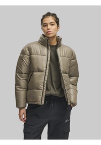 Bež puffermantel kõrge kaelusega, tikandiga disain, tõmbluku sulgemine ja mustad aktsendid. Kantud tumeda särgi ja pükste peal.