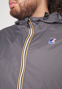 Personne barbu portant une veste K-Way grise avec une fermeture éclair rayée multicolore et un cordon de capuche ajustable.