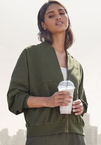 Olive green zippered bomber jacket with dropped shoulders, ribbed cuffs, and a textured fabric. Model holds a drink in a clear cup.