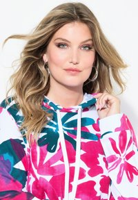 Barevná mikina s květinovým vzorem v růžové, tyrkysové a bílé barvě, s kapucí na šňůrku a jemnou texturou. Model drží kapuci.