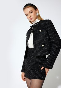 Veste en tweed noire avec des rayures argentées, design croisé, et accents en perles, associée à une mini jupe assortie et à une blouse blanche.