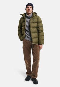 Giacca puffer verde oliva con cappuccio, maglione grigio a collo alto, pantaloni di velluto a coste marrone chiaro e stivaletti neri.