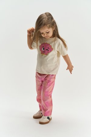 Niña joven con camiseta crema con un gráfico de una concha rosa guiñando un ojo, pantalones de cuadros rosados y zapatillas beige, mirando hacia abajo y tocándose el cabello.