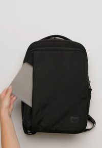 Mochila de tela negra con un diseño elegante, bolsillo delantero con cremallera y parche de marca visible. Una mano está introduciendo una funda gris para portátil en ella.