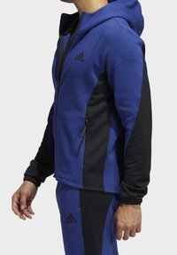 Sweat à capuche zippé bleu et noir avec une finition texturée, présentant une coupe ajustée, une capuche et des poches zippées, souligné par un logo Adidas.