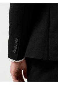 Manga de chaqueta negra con cinco botones negros. Textura suave con un corte ajustado. Puño de camisa blanca visible debajo de la chaqueta.