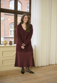 Maglione borgogna a maglia con scollo a V, abbinato a una gonna di raso borgogna e decolletè neri a punta. Pavimento in legno naturale e tende leggere.