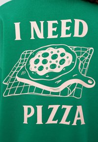 Πράσινο μπλουζάκι με λευκό κείμενο που γράφει «ΧΡΕΙΑΖΟΜΑΙ ΠΙΤΣΑ» μαζί με γραφικό μιας πίτσας σε ξυλάκι και σχέδιο με πλέγμα.