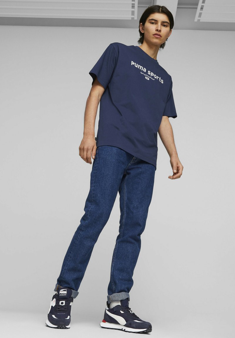 Marineblå t-skjorte med korte ermer med "Puma Sports" logo, kombinert med mørkeblå denimjeans og svarte joggesko med hvite detaljer.