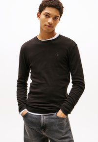 Junger Mann mit lockigem Haar, trägt ein schwarzes Langarmshirt über einem weißen Unterhemd und grauen Jeans, H�ände in den Taschen, vor einem schlichten Hintergrund.