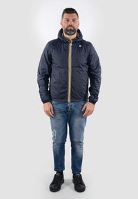 Giacca a vento blu navy con zip frontale e cappuccio, caratterizzata da strisce gialle a contrasto, jeans in denim neri e sneakers nere.