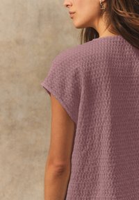 Top a maniche corte, testurizzato, in un tenue color mauve, con una vestibilità ampia e orlo leggermente curvo, mostrato da dietro con uno sfondo uniforme.