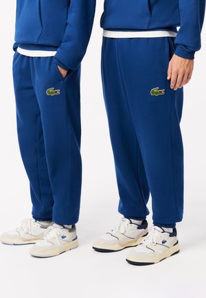 Blauwe fleece joggingbroek met elastische boorden en zakken, met een groen geborduurd krokodillenlogo op het linkerbeen, gecombineerd met witte sneakers.