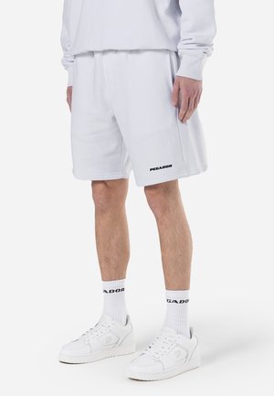 Persona que lleva pantalones cortos deportivos blancos, calcetines blancos a media pierna con texto negro y zapatillas blancas, de pie frente a un fondo liso.