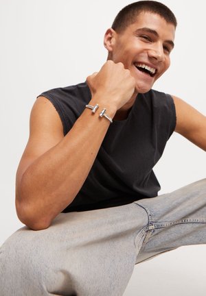 Hombre sonriente con una camiseta sin mangas negra y jeans claros, mostrando una pulsera de brazalete plateada de extremos abiertos en su muñeca levantada.