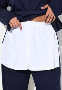 Blusa blanca de botones con cintura elástica y aberturas laterales, llevada sobre pantalones azul marino. La tela es suave; el diseño presenta un corte relajado.