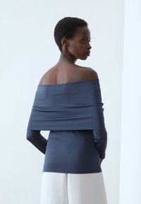 Jūras spēku off-the-shoulder tops ar gariem piedurkņu, gludu tekstūru un pieguļošu dizainu. Apskatā mugurskatu izceļ maigu drapējumu pāri pleciem.