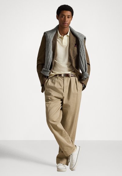 Καφέ σακάκι πάνω από ανοιχτό πόλο, γκρι πλεκτό πουλόβερ περασμένο, άνετο μπεζ παντελόνι και λευκά αθλητικά παπούτσια, δημιουργώντας μια χαλαρή αλλά καλλωπισμένη εμφάνιση.