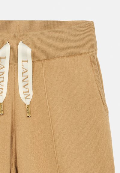 Béžové tepláky mají ribovaný elastický pas s šňůrkami, zdobené zlatými špičkami. Hladký materiál s bočními kapsami.