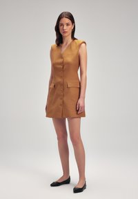 Vestido sin mangas mostaza con escote en V, que presenta un cierre de botones, dos bolsillos y un corte entallado. Combinado con zapatos planos negros.