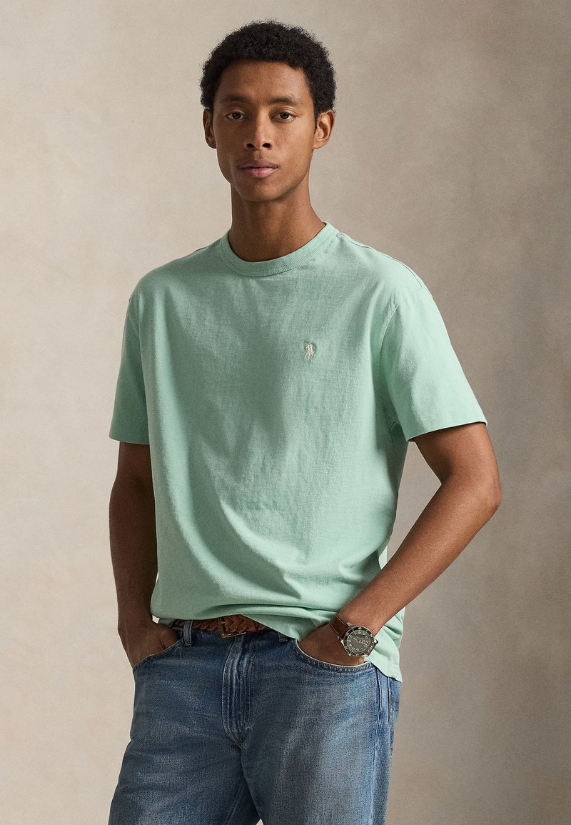 T-shirt de algodão verde claro com gola redonda, corte descontraído, com um pequeno logotipo no peito. Combinada com jeans azul desbotado.