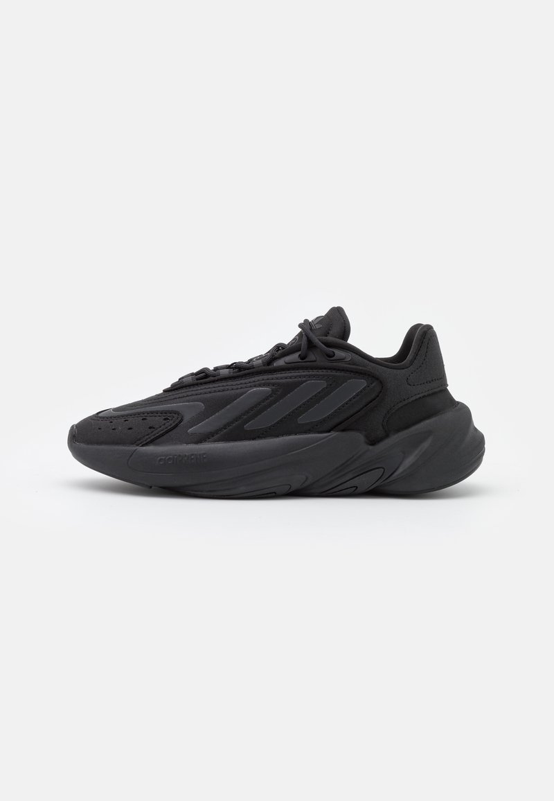 Zapatilla deportiva negra con parte superior de textil suave que presenta elementos de diseño en capas, media suela redondeada y suela de goma texturizada.