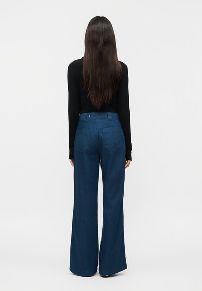 Femme aux longs cheveux bruns portant un haut noir à manches longues et un jean bleu taille haute à jambes larges, debout de dos devant un fond uni.