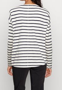 Persoon in een wit overhemd met lange mouwen en zwarte horizontale strepen, en een zwarte broek, van achteraf gezien tegen een effen achtergrond.