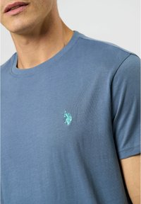 Světlemodré bavlněné tričko s kulatým výstřihem, krátkými rukávy a malým vyšitým logem v mátově zelené barvě na hrudi.