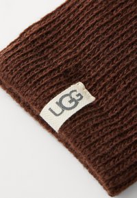 Brun strikket lue med en teksturert overflate og synlig UGG-logo i kremfarge, som viser myke, tykke fibre.