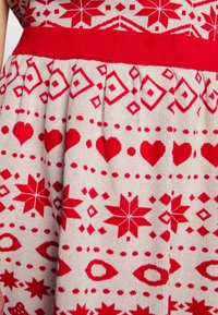 Vestido de malha com fundo creme, apresentando padrões geométricos vermelhos, corações e estrelas; inclui uma faixa vermelha na cintura; textura suave.