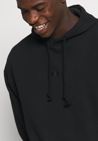 Μαύρη κουκούλα φτιαγμένη από ύφασμα μείγματος βαμβακιού, με κορδόνι, τσέπη καγκουρό και διακριτική λεπτομέρεια λογότυπου στο στήθος.