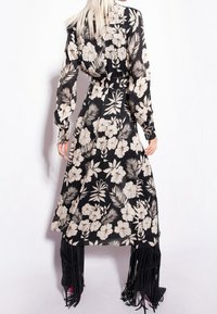 Kobieta w czarnej sukience midi z beżowym kwiatowym wzorem i długimi rękawami, w połączeniu z czarnymi, frędzlowymi szpilkami, stojąca na białym tle.