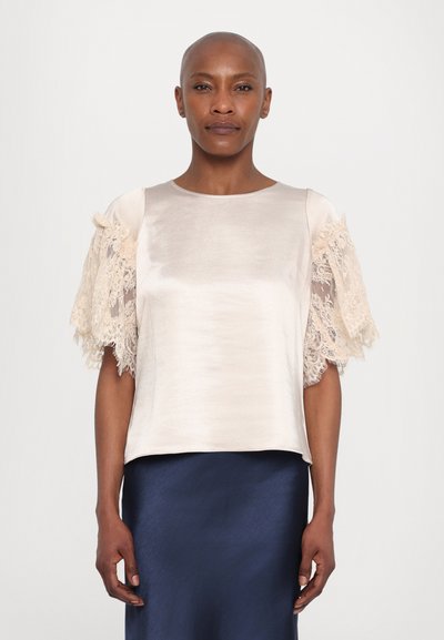 Haut en satin crème à manches courtes avec des manches bordées de dentelle, encolure arrondie et ourlet droit. Porté avec une jupe bleu marine.