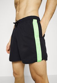 Shorts da nuoto neri con fascia elastica in vita, cordoncino e una striscia logo verde a contrasto lungo il lato. Il materiale è leggero e liscio.