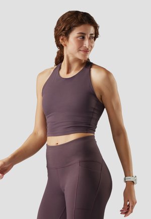 Frau in lila sportlichem Crop-Top und Leggings, die seitlich schaut und an ihrem linken Handgelenk eine Fitnessuhr trägt.