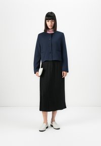 Femme portant une veste boutonnée bleu marine, un col roulé rose, une jupe midi plissée noire, des mocassins blancs et tenant une petite pochette blanche.