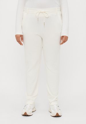Light cream sweatpants with a drawstring waistband, side pockets, and a tapered fit. Paired with white athletic shoes. Soft fabric texture.