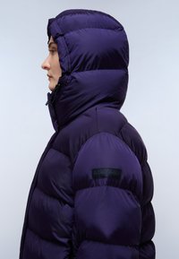 Fioletowa kurtka puchowa z wysokim kołnierzem i kapturem, o błyszczącej fakturze i pikowanym wzorze. Naszywka z logo na lewym rękawie.