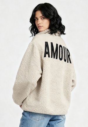 Veste en polaire beige avec surface texturée, comportant un panneau lisse contrastant au dos. "AMOUR" est brodé en noir de manière bien visible.