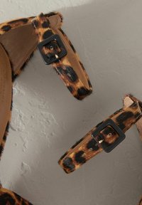 Sandalen mit Leopardenmuster und einer fellähnlichen Textur, ausgestattet mit schwarzen Schnallen und einem glatten, braunen Innenfutter, vor einem hellgrauen Hintergrund.