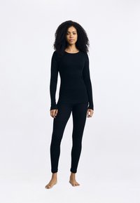 Mulher com cabelo encaracolado, descalça, vestindo uma blusa preta de mangas longas justa e leggings pretas, contra um fundo branco simples.