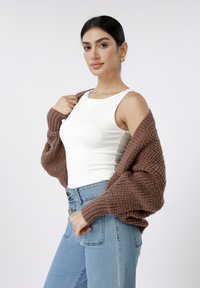 Καφέ πλεκτή ζακέτα πάνω από λευκό αμάνικο τοπ, σε συνδυασμό με ψηλόμεσο μπλε τζιν με πέντε κουμπιά και εφαρμοστή γραμμή.