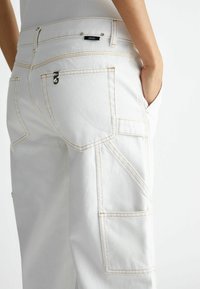 Białe spodnie cargo z kontrastującymi żółtymi szwami, z wieloma kieszeniami i metką na tylnej części pasa. Gładka tekstura denimu.