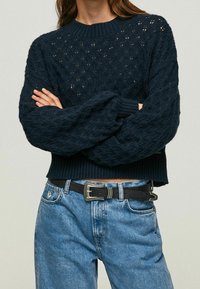 Granatowy sweter dziany z wzorem diamentowym, z prążkowanym dekoltem i mankietami, w połączeniu z jasnoniebieskimi dżinsami i czarnym paskiem z metalową klamrą.