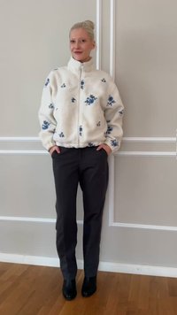 Γυναίκα στέκεται μπροστά σε μπεζ τοίχο φορώντας λευκό φλις μπουφάν με φερμουάρ και μπλε λουλουδάτα σχέδια, μαύρο παντελόνι και μαύρα μποτάκια μέχρι τους αστραγάλους, με τα χέρια στις τσέπες.