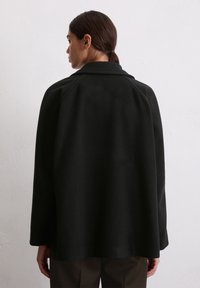 Tumedate juuksed tagasi seotud naine kannab lahtist musta mantlit ja tumedaid pruune pükse, seistes ühetoonilise heledat värvi seina vastu, selg kaamera poole.